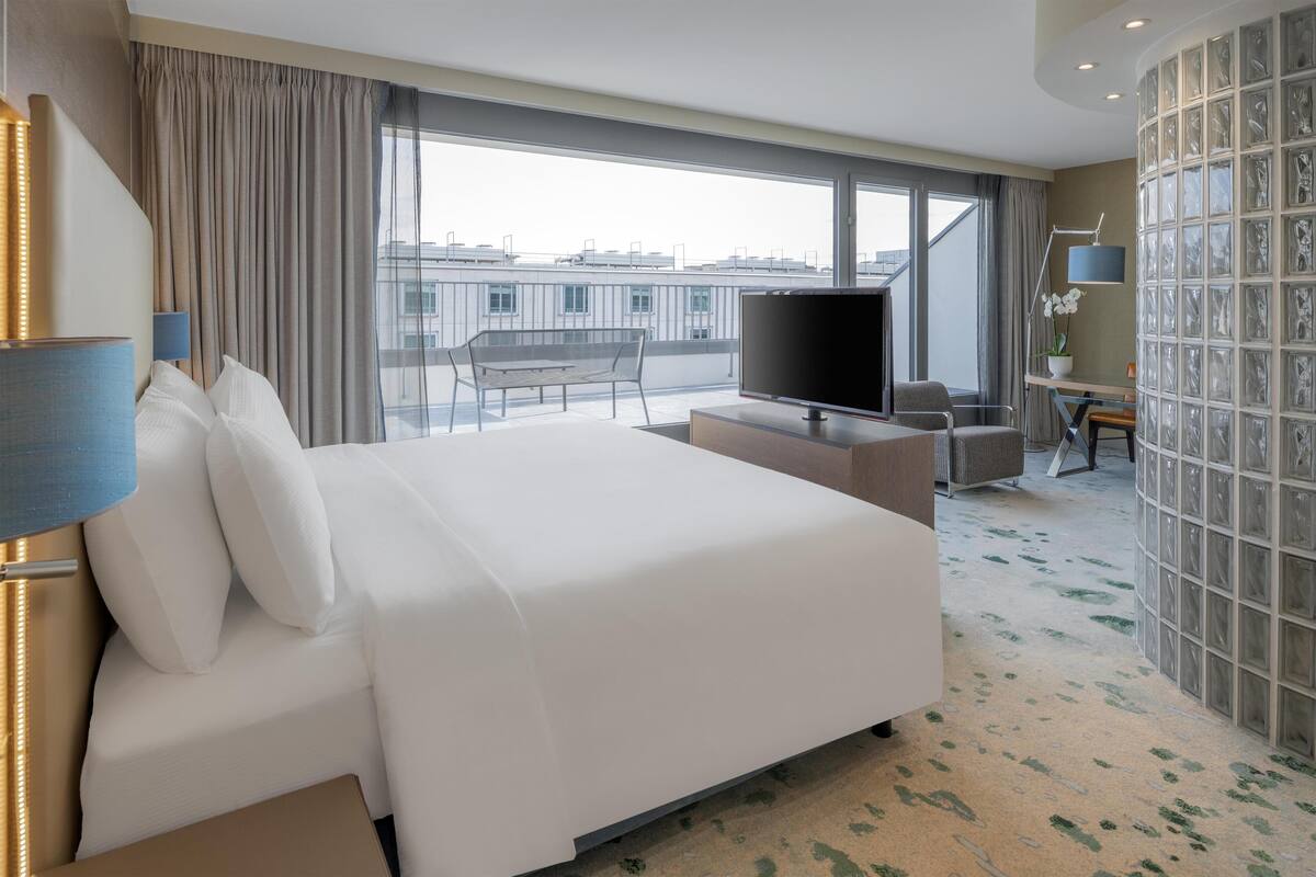 A modern hotel room with a large bed, a flat-screen TV, a desk, and a view of the cityscape through large windows.