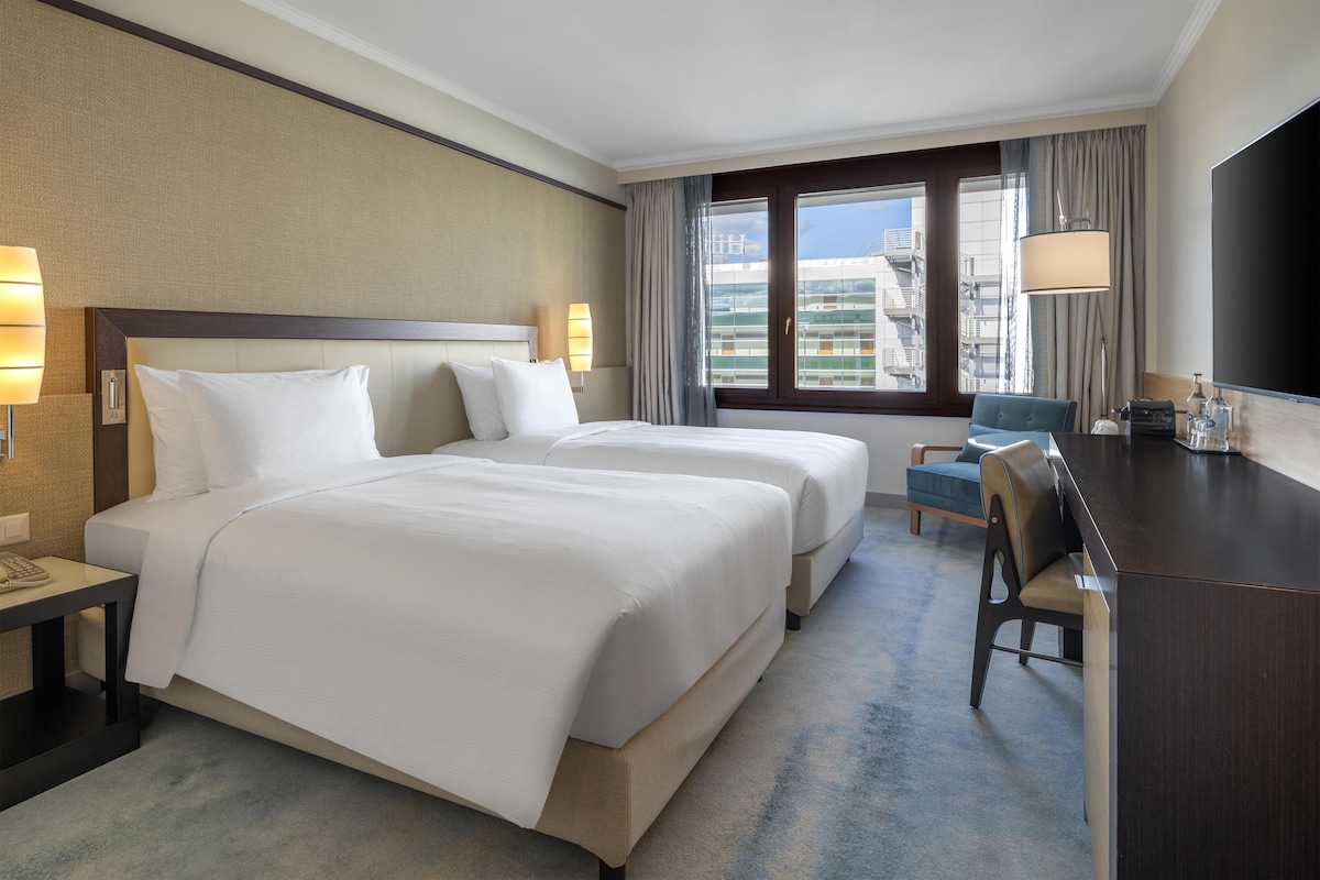 A hotel room with two beds, a desk, a chair, and a large window with a view of buildings.