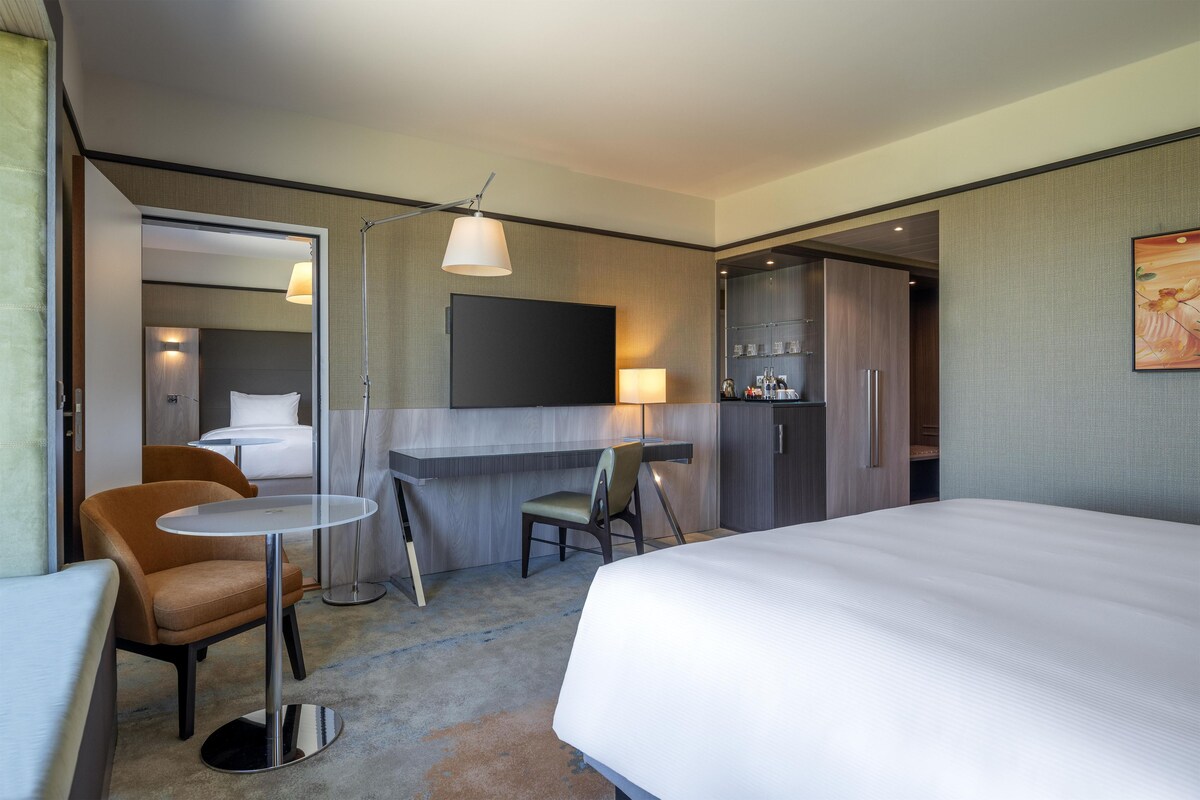 A hotel room with a bed, a desk, a chair, a small table, a sofa, a TV, and a wardrobe.