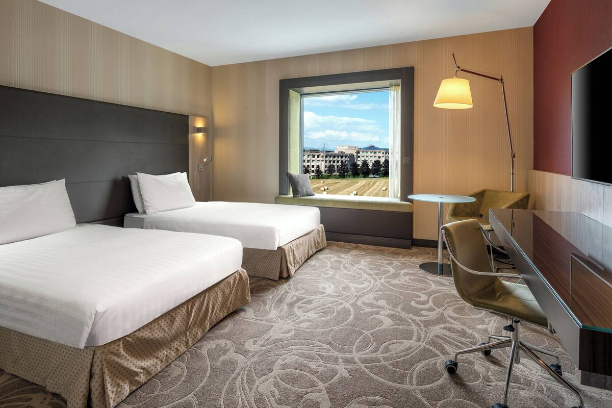 A hotel room with two beds, a desk, a chair, a lamp, and a large window with a view.