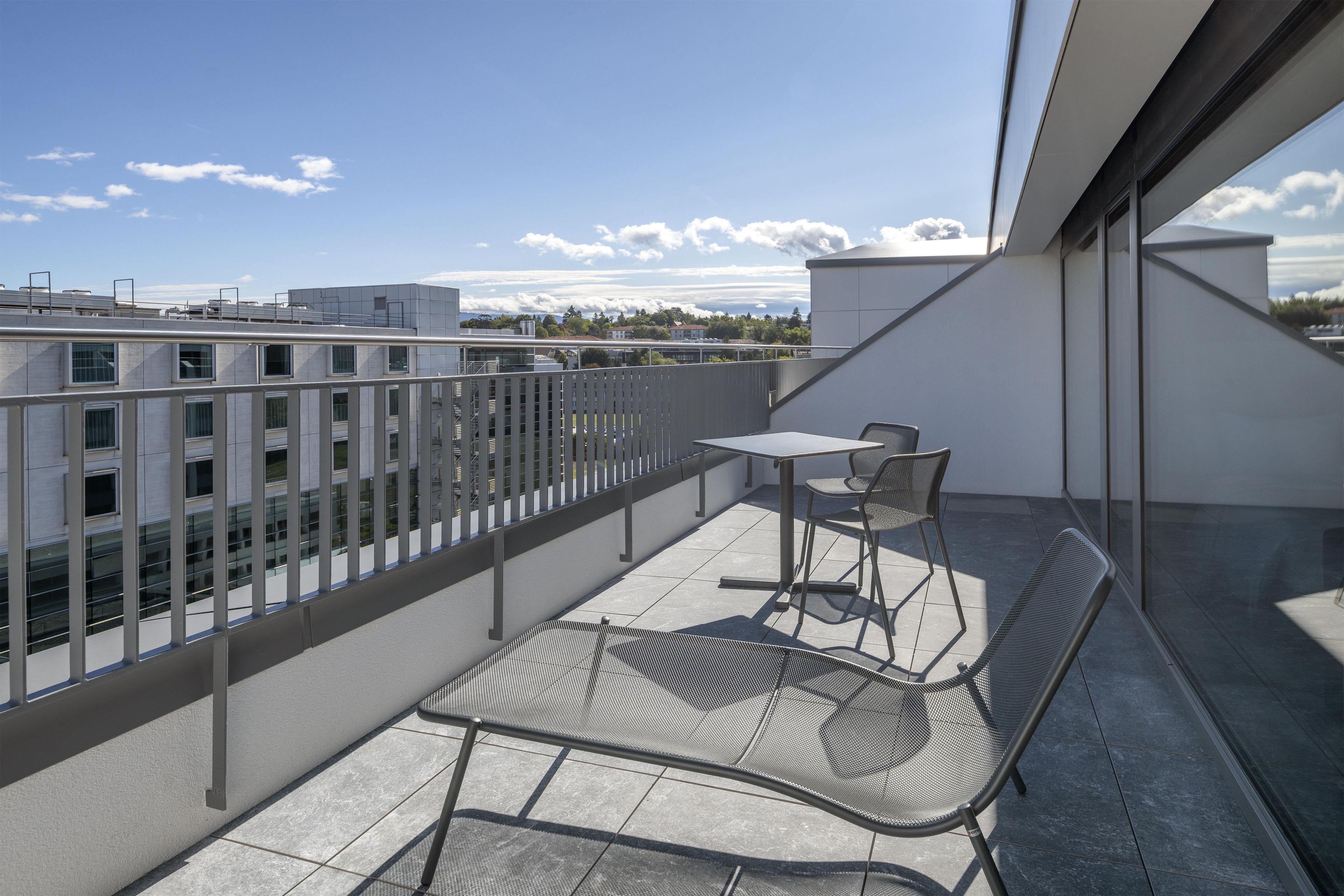 A balcony with a table and two chairs, a metal railing, and a view of buildings and trees.