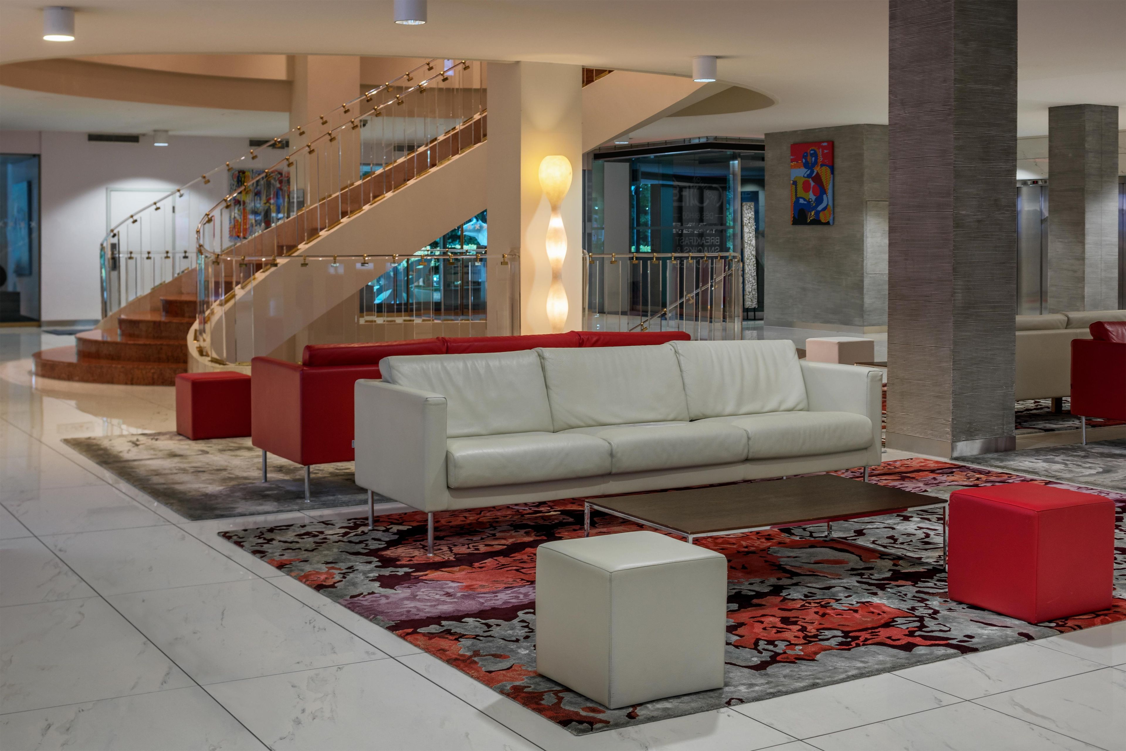 A modern lobby with a white leather sofa, red accent chairs, a patterned rug, and a staircase with glass railings.