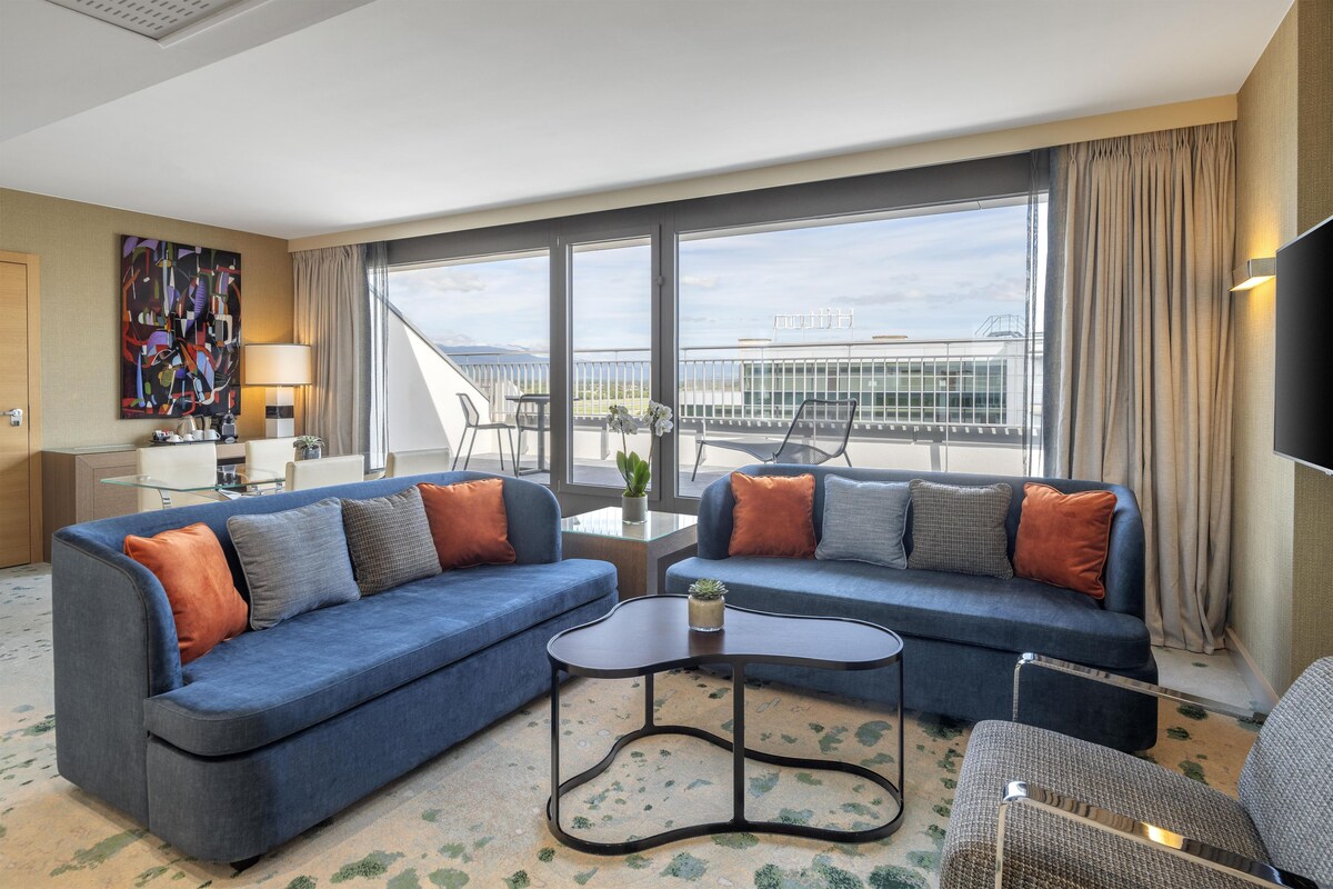 A modern hotel room with a blue sofa set, a small coffee table, and a view of the cityscape through large windows.