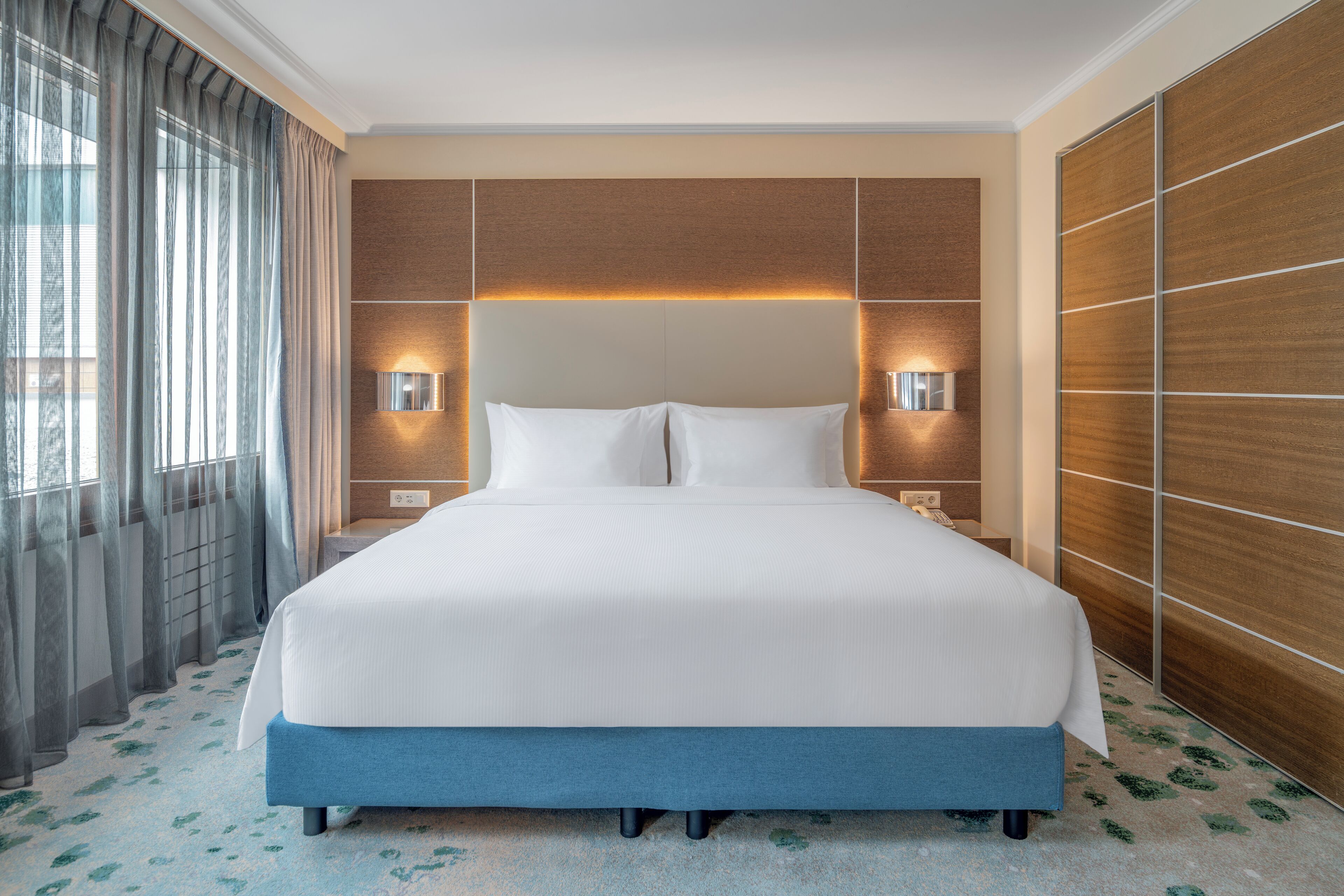 A hotel room with a large bed, wooden headboard, two bedside lamps, and a view through a window with curtains.