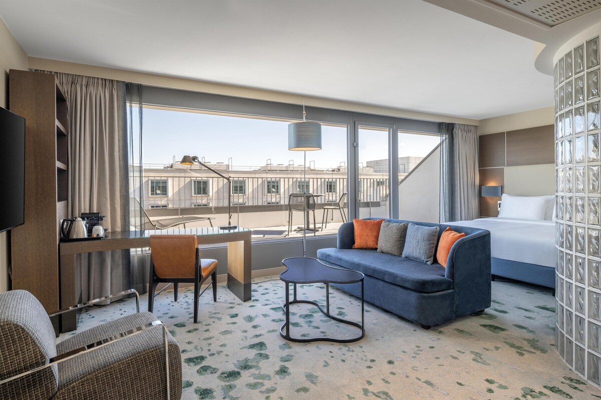 A hotel room with a bed, a sofa, a desk, and a chair. There is a large window with a view of buildings outside.