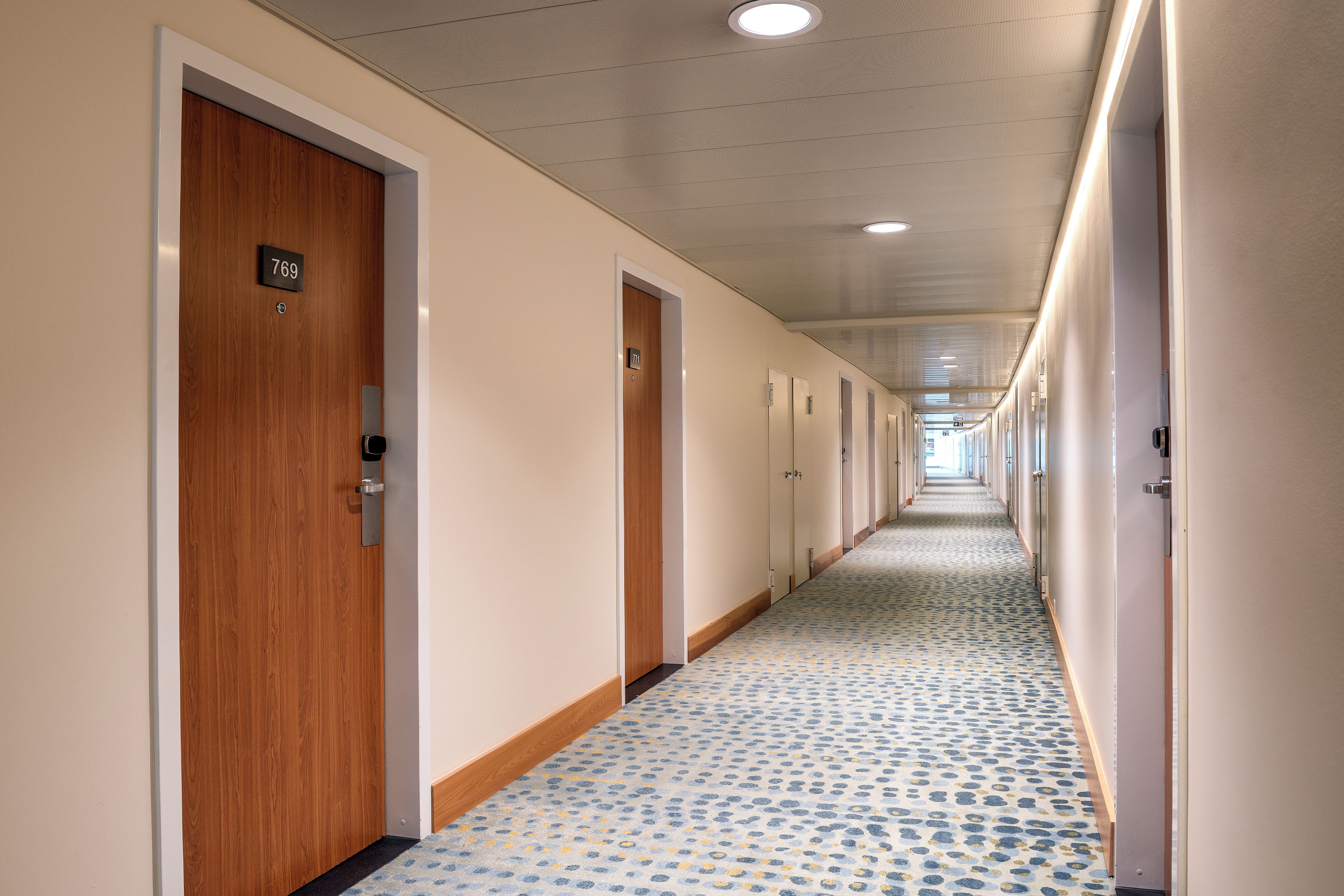 A hotel corridor with wooden doors, numbered room signs, and patterned carpet.
