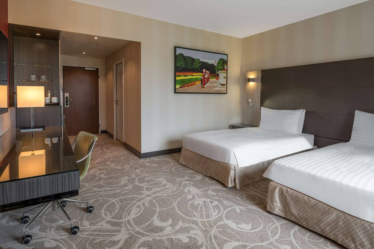 A hotel room with two beds, a desk with a lamp, a chair, and a painting on the wall.