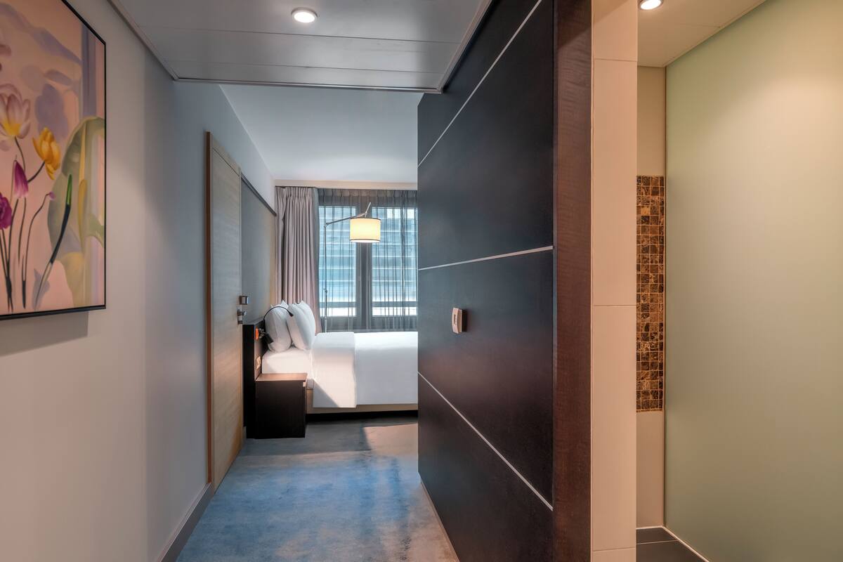 A hotel corridor with a bed, a nightstand, a lamp, and a painting on the wall.