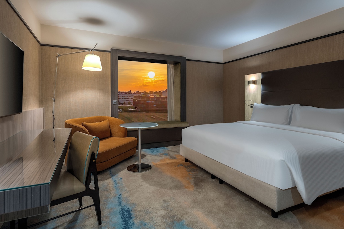 A hotel room with a bed, a chair, a small table, and a view of a sunset.