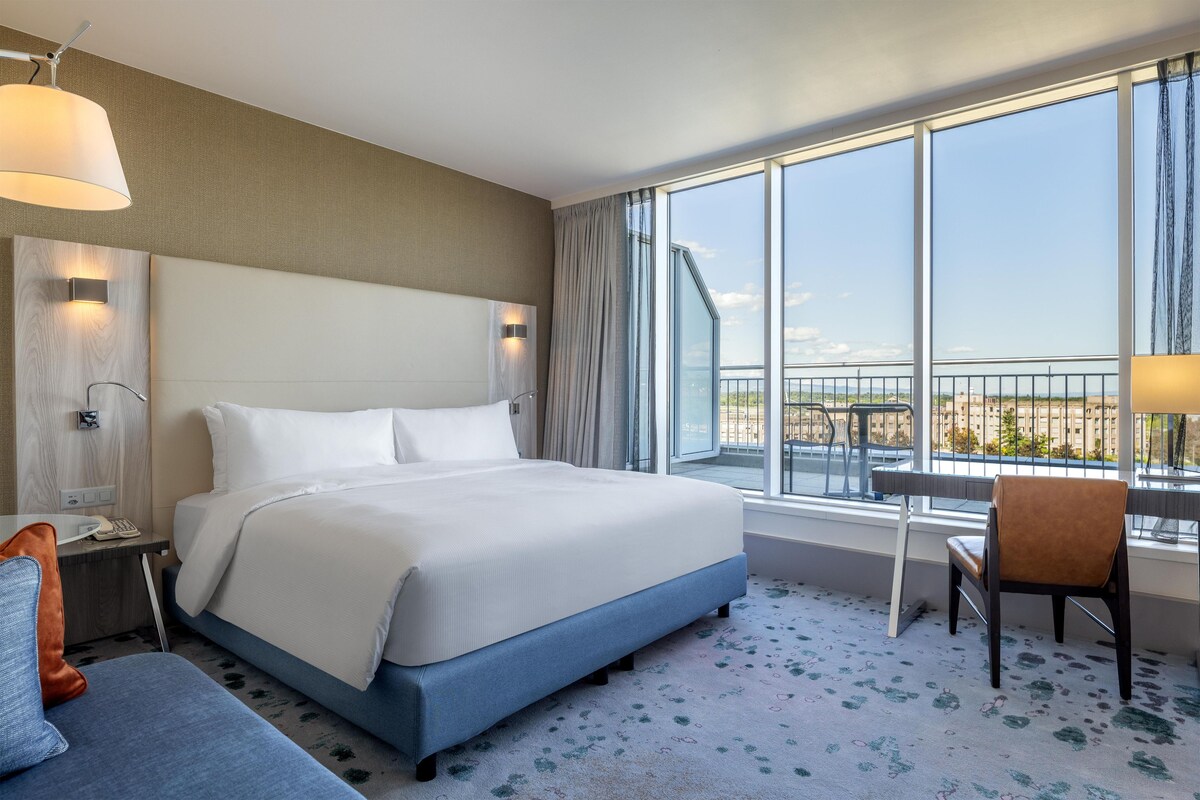 A hotel room with a large bed, a desk, a chair, a window with a view, and a ceiling fan.