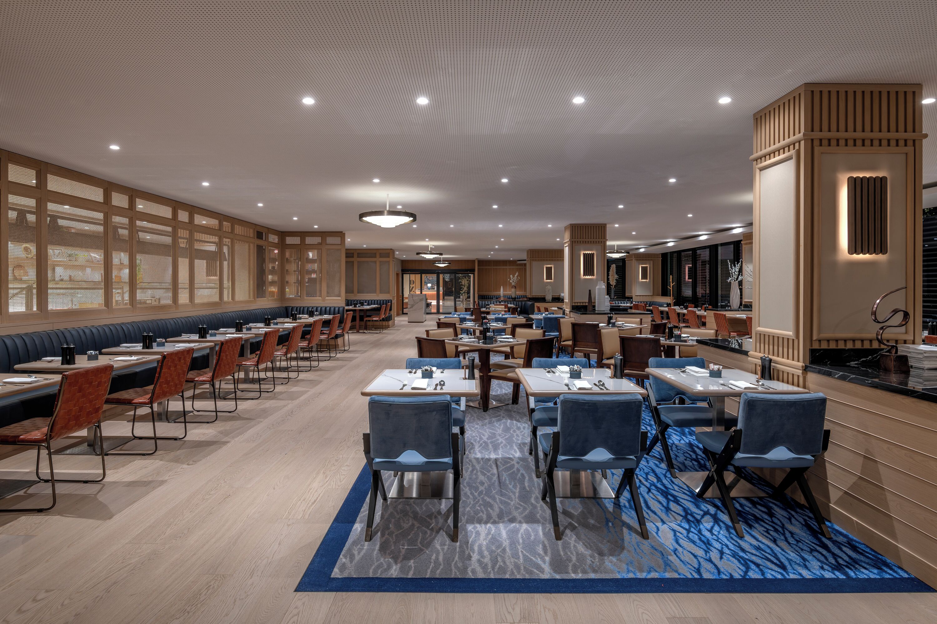 Un comedor amplio con varias mesas y sillas, alfombra estampada y un diseño moderno de techo.