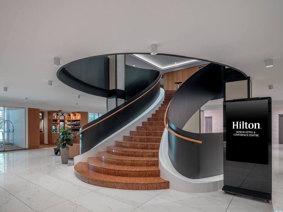 Interior moderno de hotel con una escalera curva, una zona de recepción y un amplio ventanal con vistas al exterior.