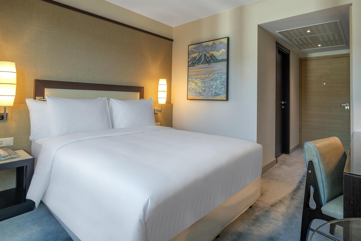 A hotel room with a large bed, bedside lamps, a painting on the wall, a door, and a chair.