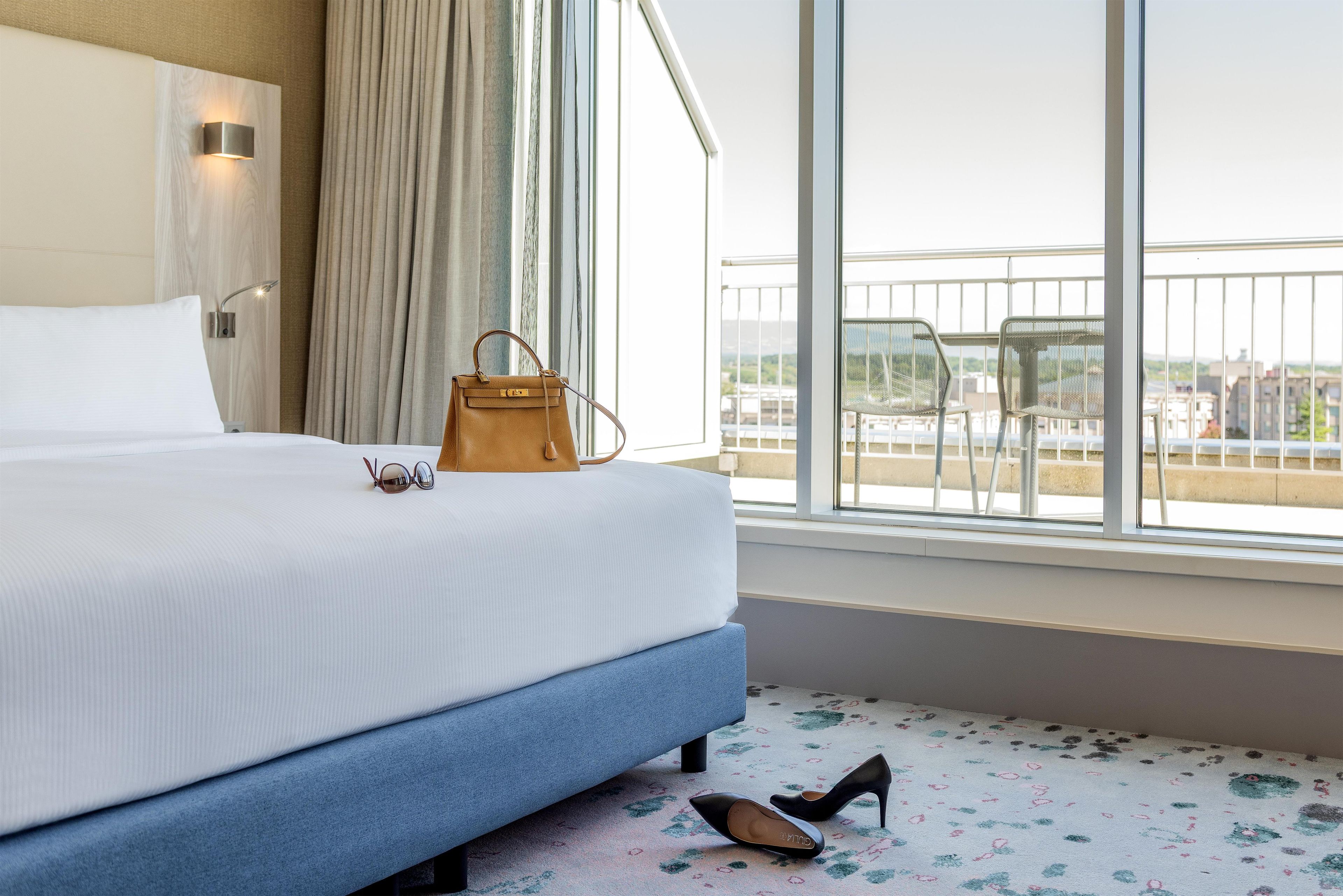 A hotel room with a bed, a window with a view of the city, a balcony with chairs, and a handbag on the bed.