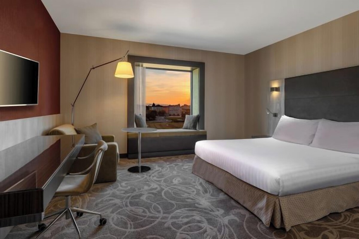 Quarto de hotel com cama, escrivaninha, cadeira e vista para a cidade ao pôr do sol.