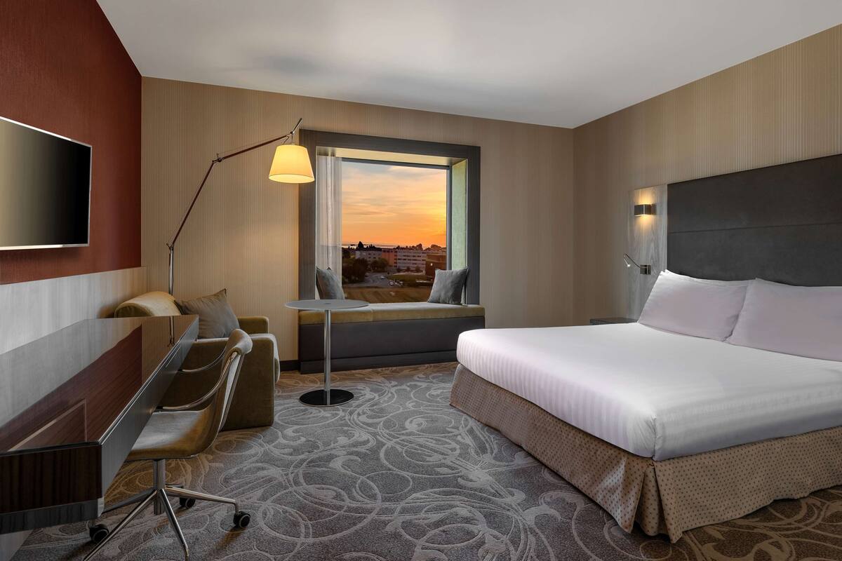 A hotel room with a bed, desk, chair, and a view of the sunset through a window.