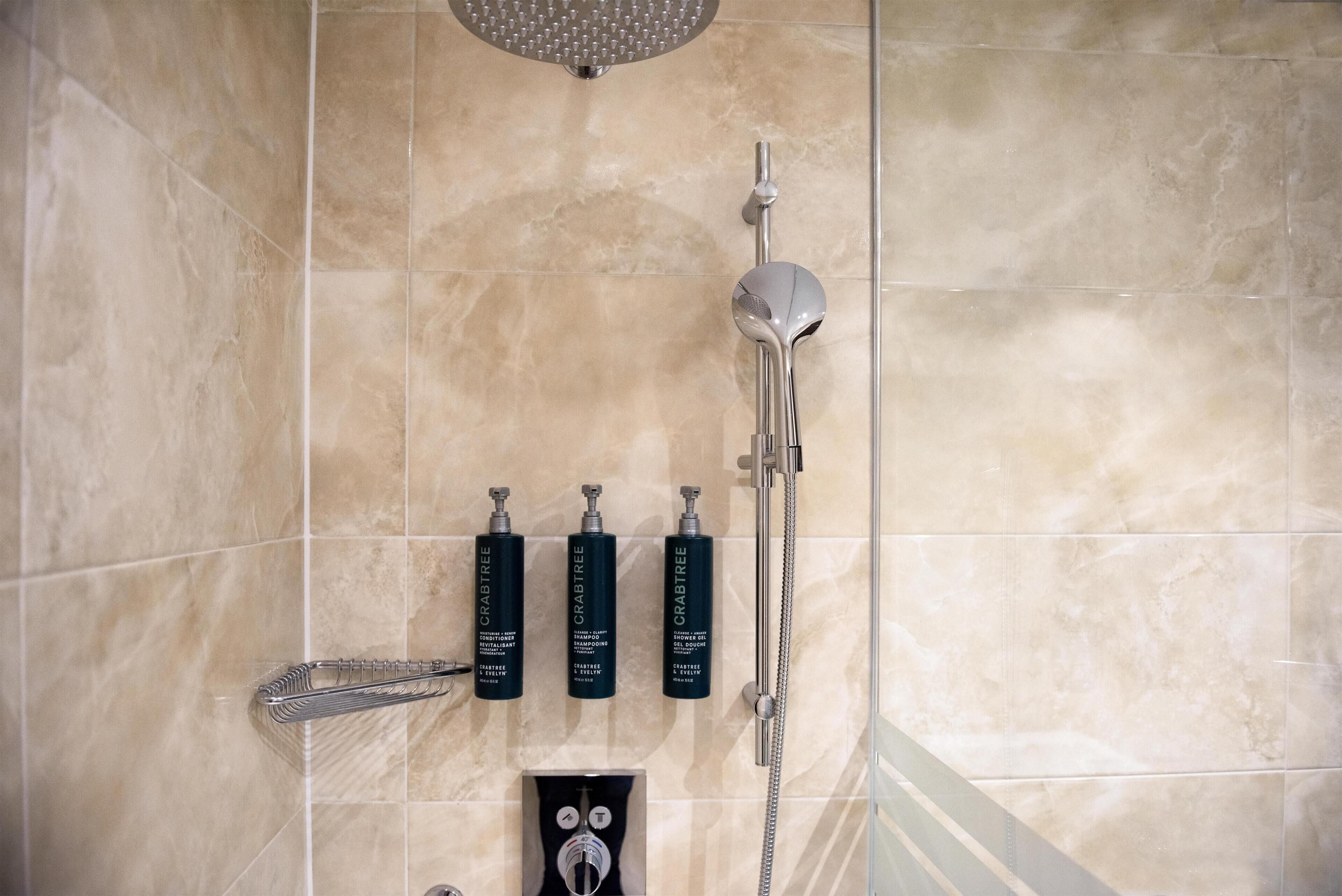 A bathroom with a shower, a hand dryer, and three bottles of Crabtree shampoo.