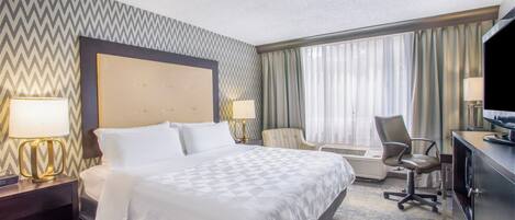 Habitación de hotel con una cama grande, dos mesitas de noche con lámparas, un escritorio con computadora, una silla y una ventana con cortinas.