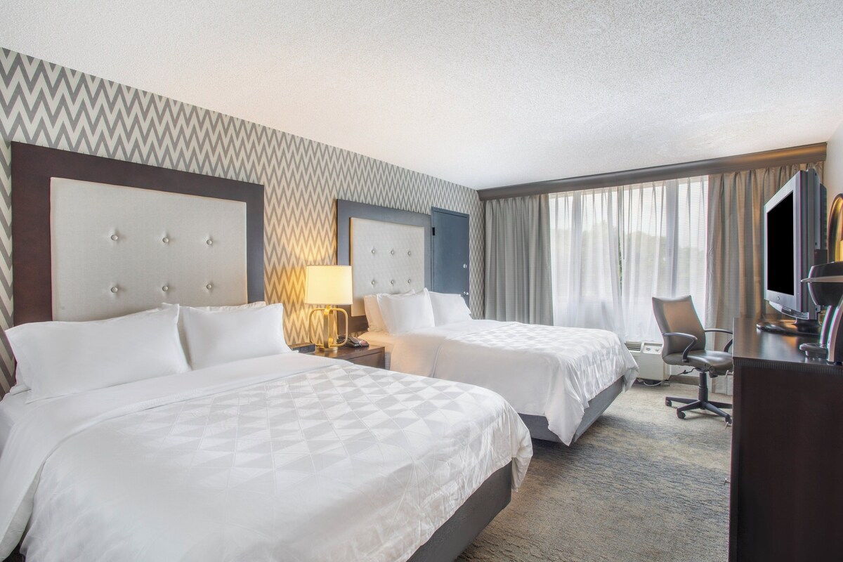 A hotel room with two beds, a desk, a chair, a TV, and a large window with curtains.