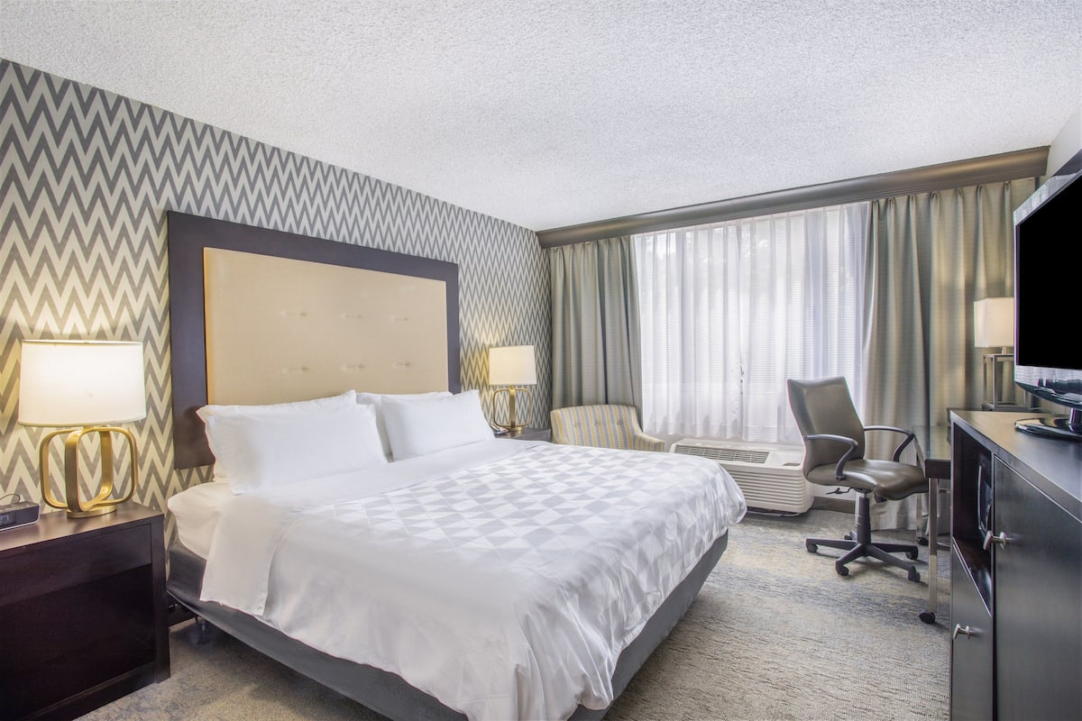 Ein Hotelzimmer mit einem großen Bett, zwei Nachttischlampen, einem Schreibtisch mit einem Computer, einem Sessel und einem Fernseher.