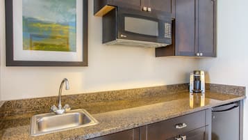 A kitchen with a granite countertop, stainless steel sink, dark wooden cabinets, a microwave, and a coffee maker.
