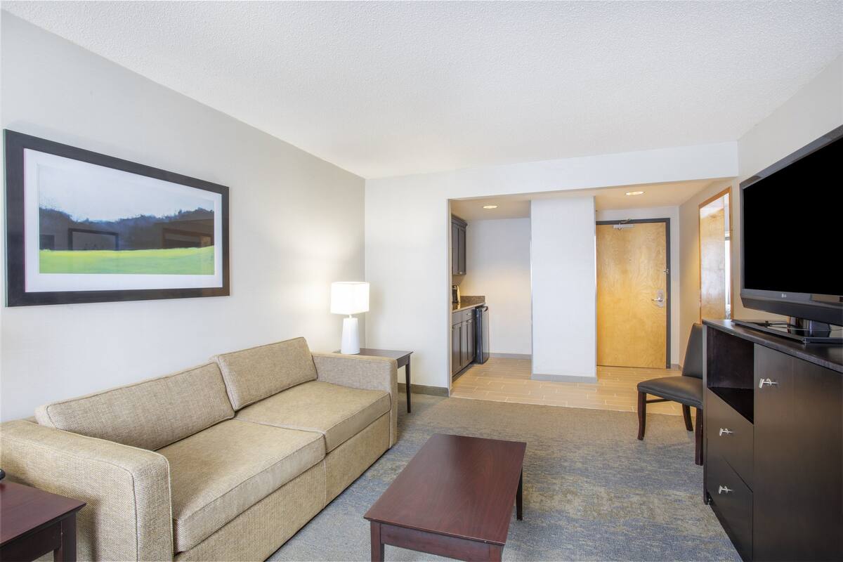 Ein Hotelzimmer mit einer Couch, einem Couchtisch, einem Fernseher, einem Schreibtisch und einem gerahmten Bild an der Wand.
