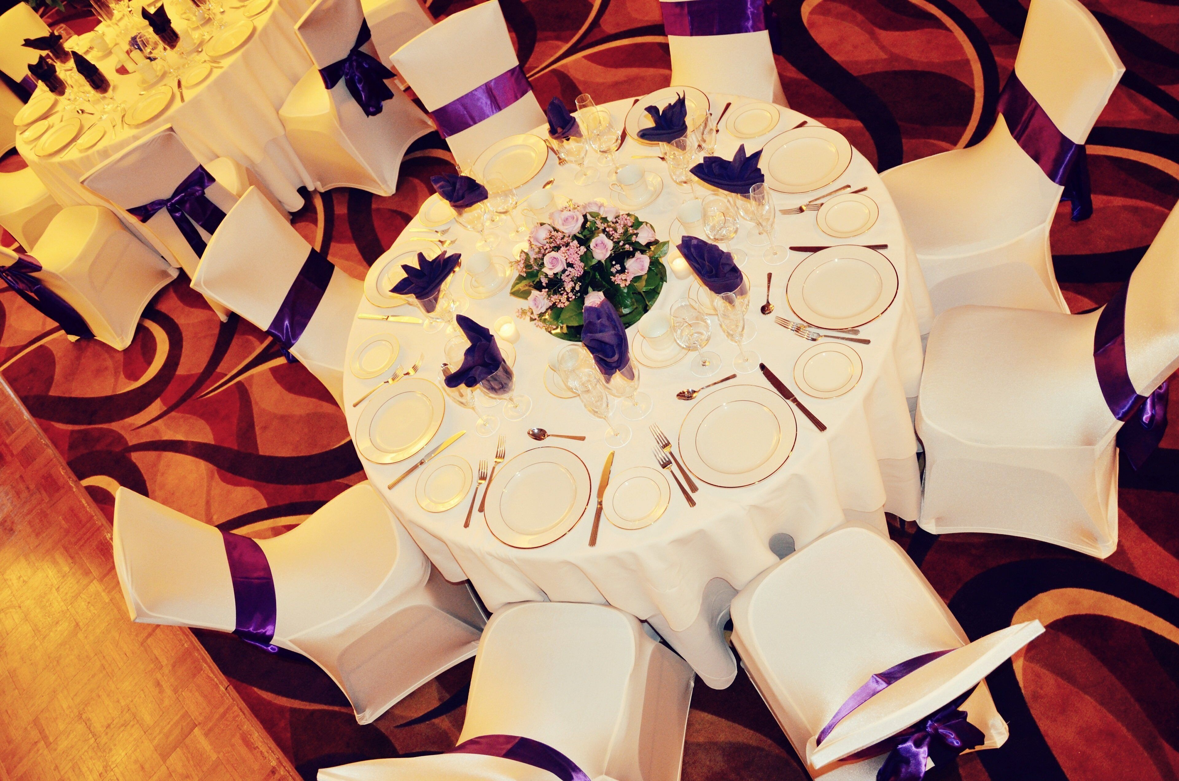 A round table with white tablecloths, purple sashes, and floral centerpieces.
