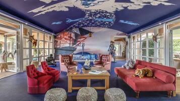 Relax in a spacious room with a high ceiling, featuring a nautical mural and comfortable seating area with a sofa, armchairs, and woven poufs.