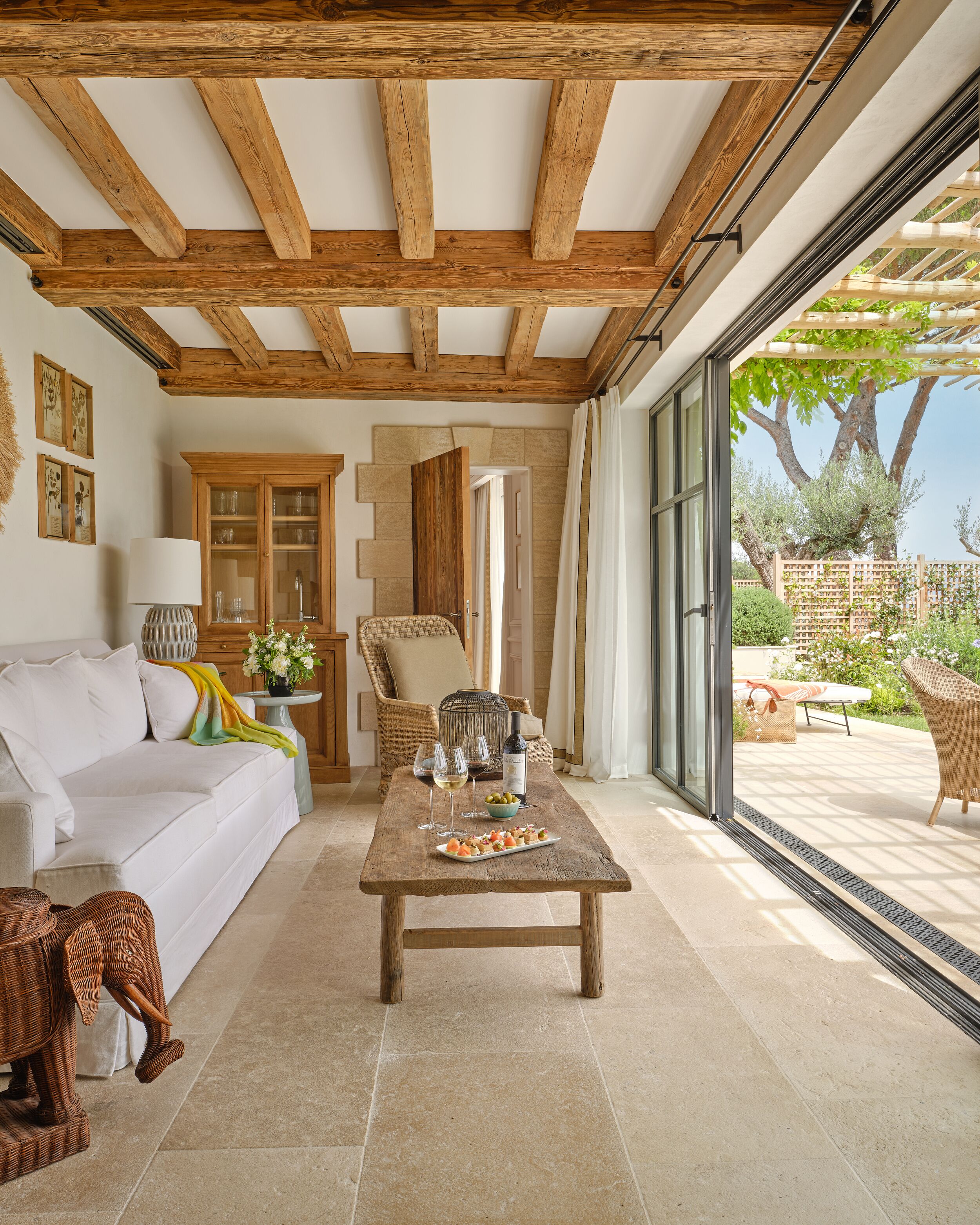 A spacious living area with a wooden ceiling, a white sofa, a dining table with a meal, and a view of the garden through large windows.