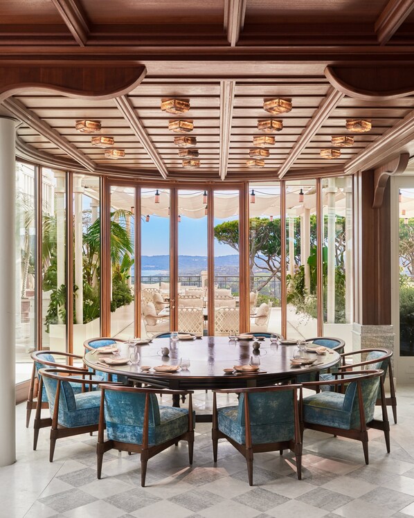 A dining area with a round table, blue upholstered chairs, and a view of the outdoors. - Airelles Château de la Messardière (Saint-Tropez)
