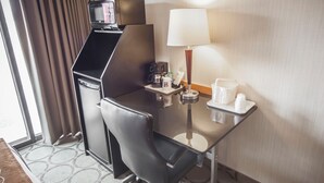 A hotel room with a compact desk, a coffee maker, a lamp, and a small table with cups and a bottle.