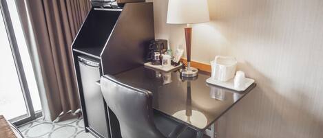 A hotel room with a compact desk, a coffee maker, a lamp, and a small table with cups and a bottle.