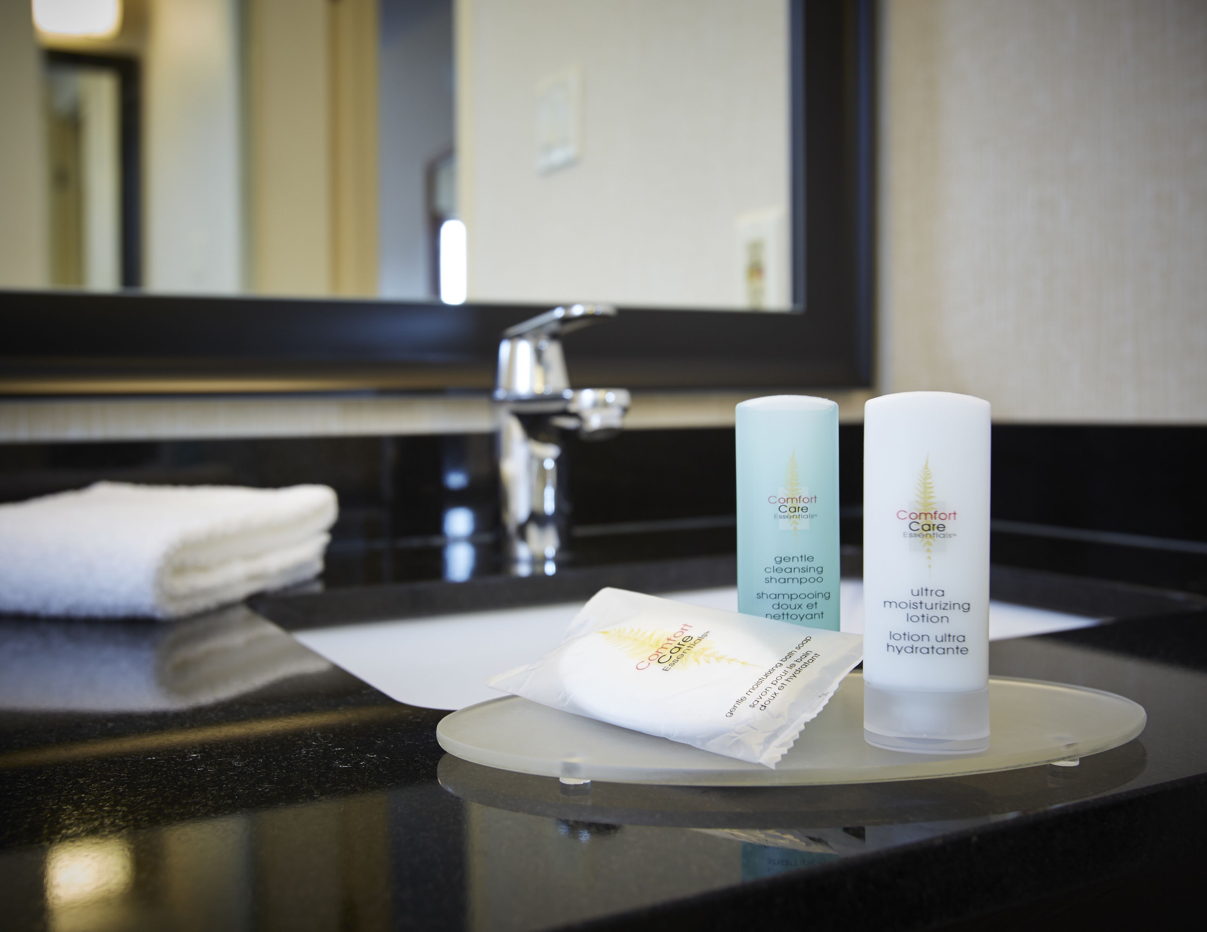 A bathroom vanity with a white towel, two bottles of Comfort Care Essentials shampoo and lotion, and a mirror above.