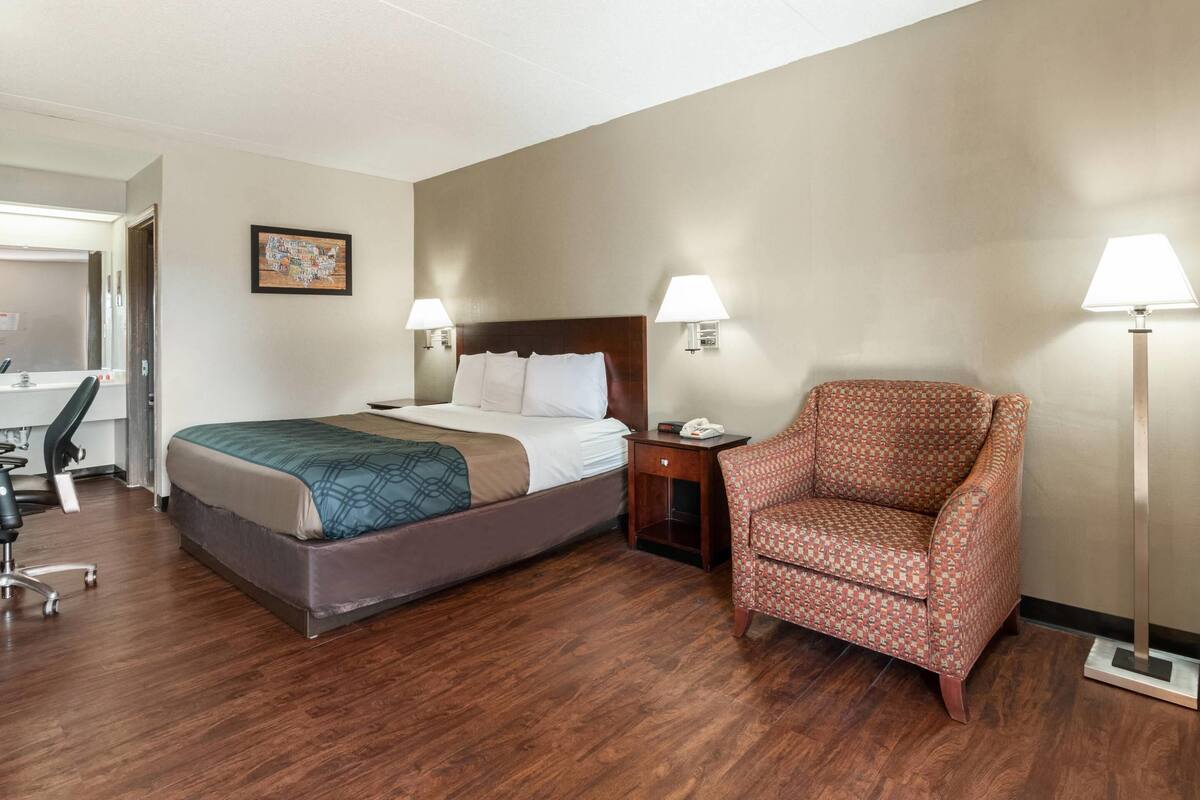 A hotel room with a bed, a chair, a desk, and a lamp.