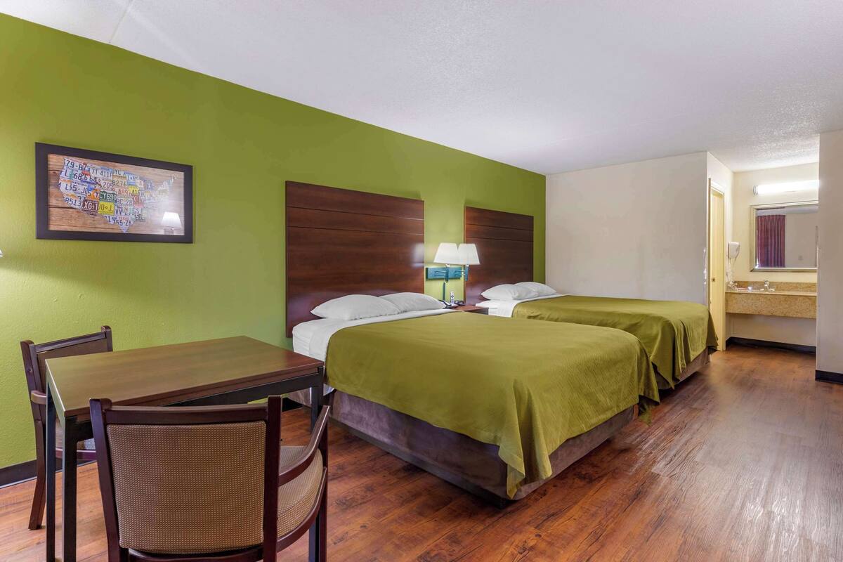 A hotel room with a wooden table, two single beds with green bedspreads, a headboard, a nightstand with a lamp, and a framed map on the wall.