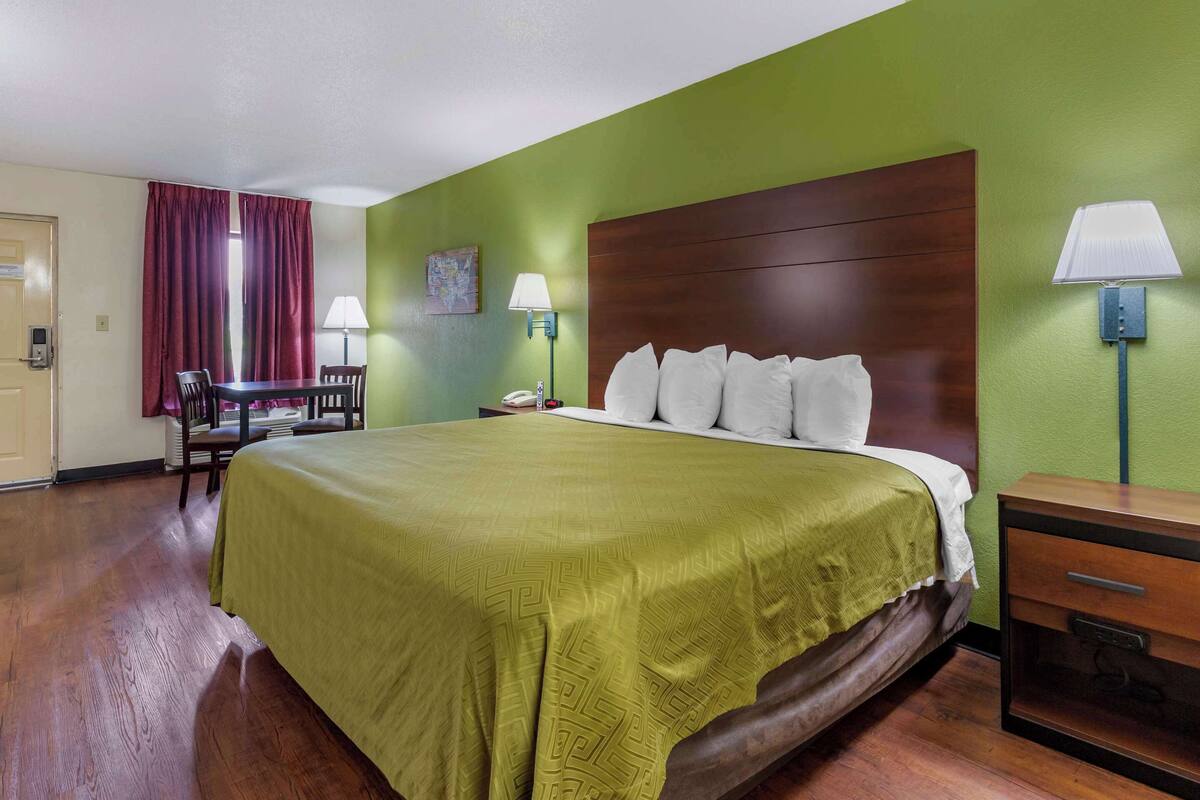 A hotel room with a large bed, a wooden nightstand, a desk with a chair, and a green wall.