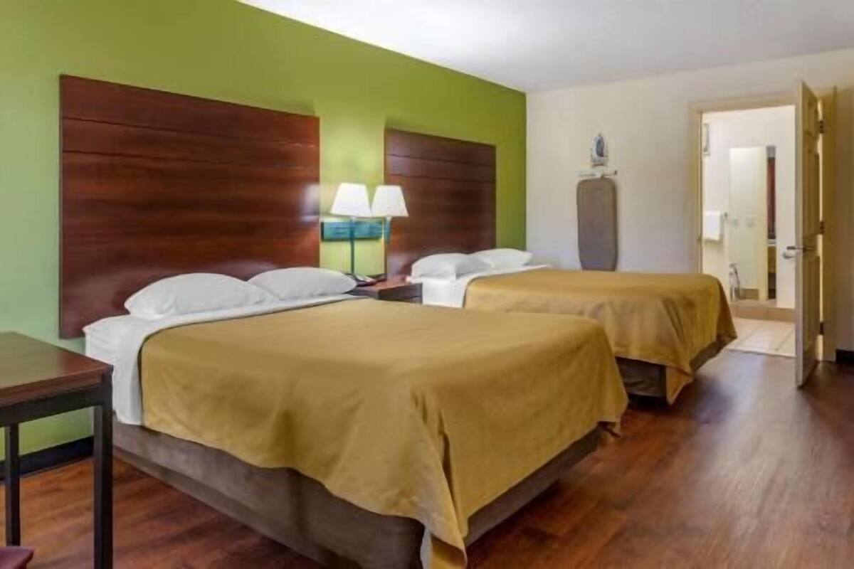 A hotel room with two beds, wooden headboards, a nightstand with a lamp, and a door leading to another room.