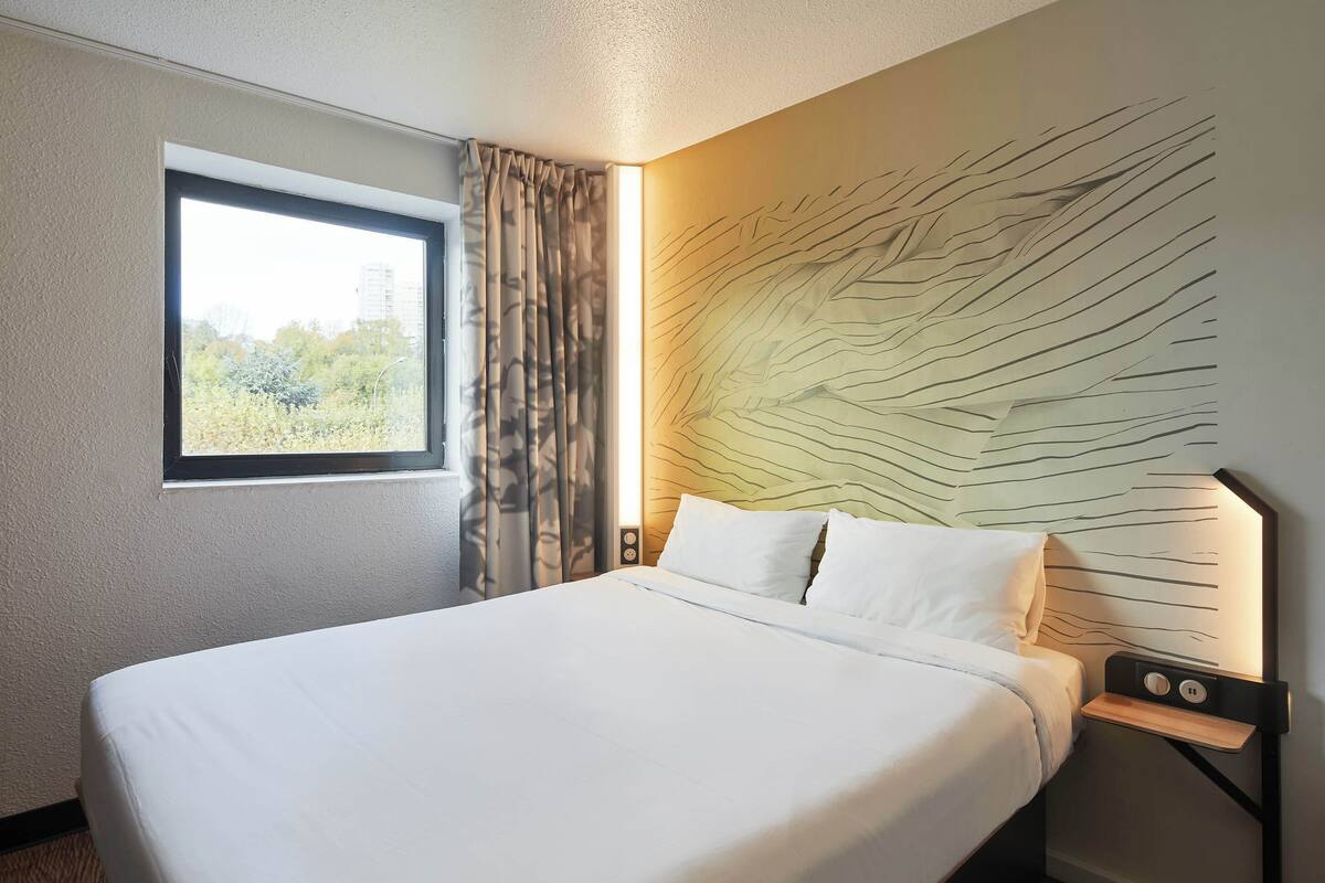 A hotel room with a large bed, a window with curtains, a wall-mounted light, and a nightstand with a lamp.