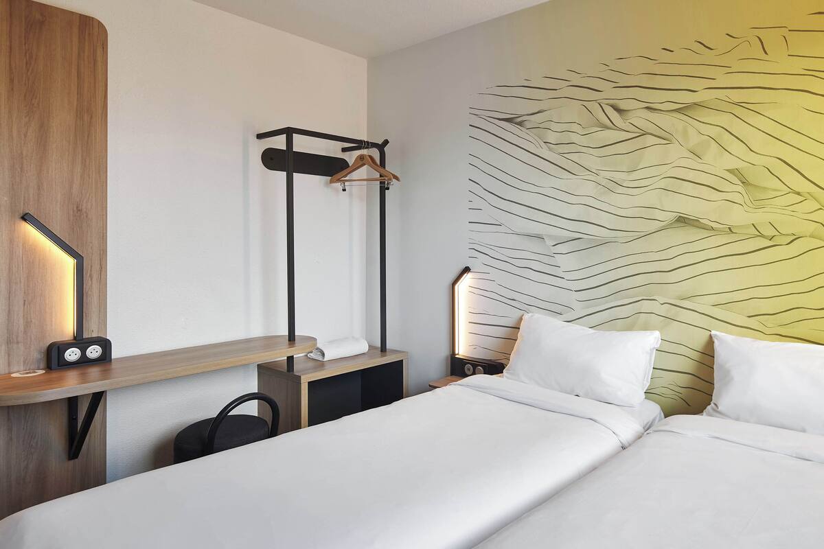 A hotel room with a bed, bedside table, wall-mounted lamp, and a hanging rack.