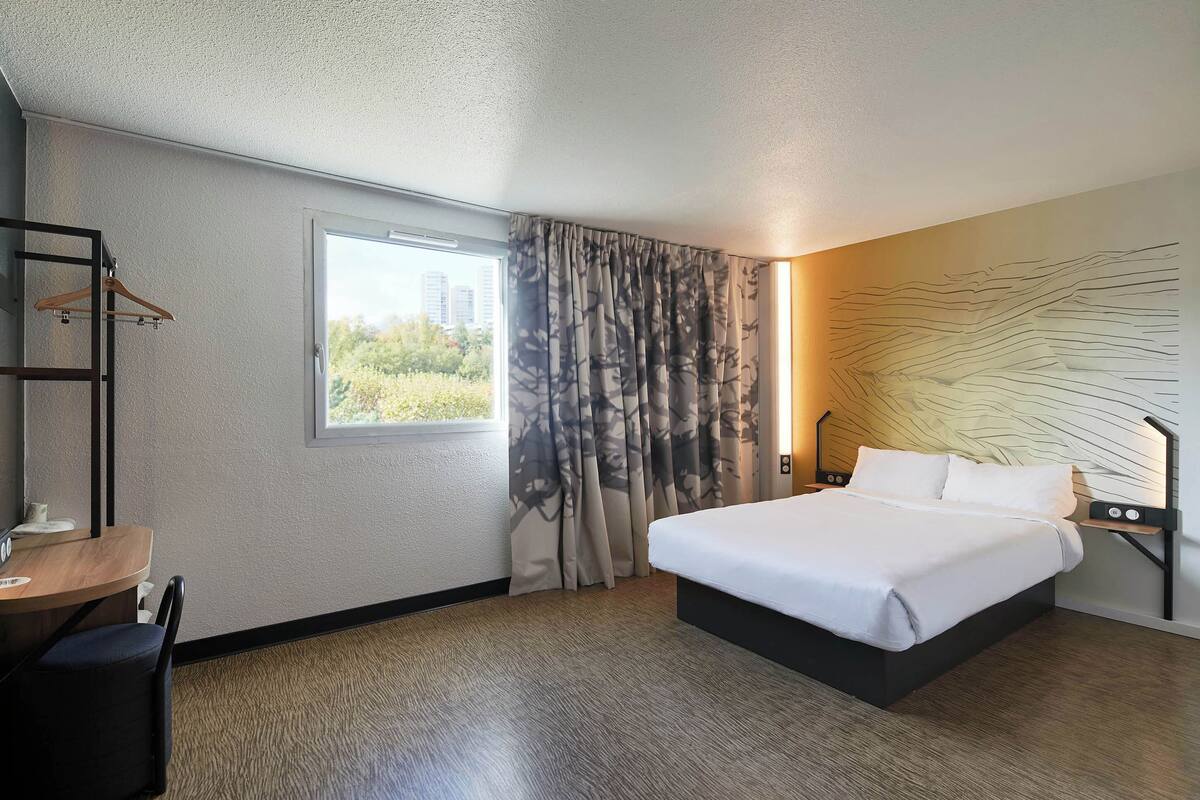 A hotel room with a bed, a desk, a chair, a window with curtains, and a wall with a textured design.