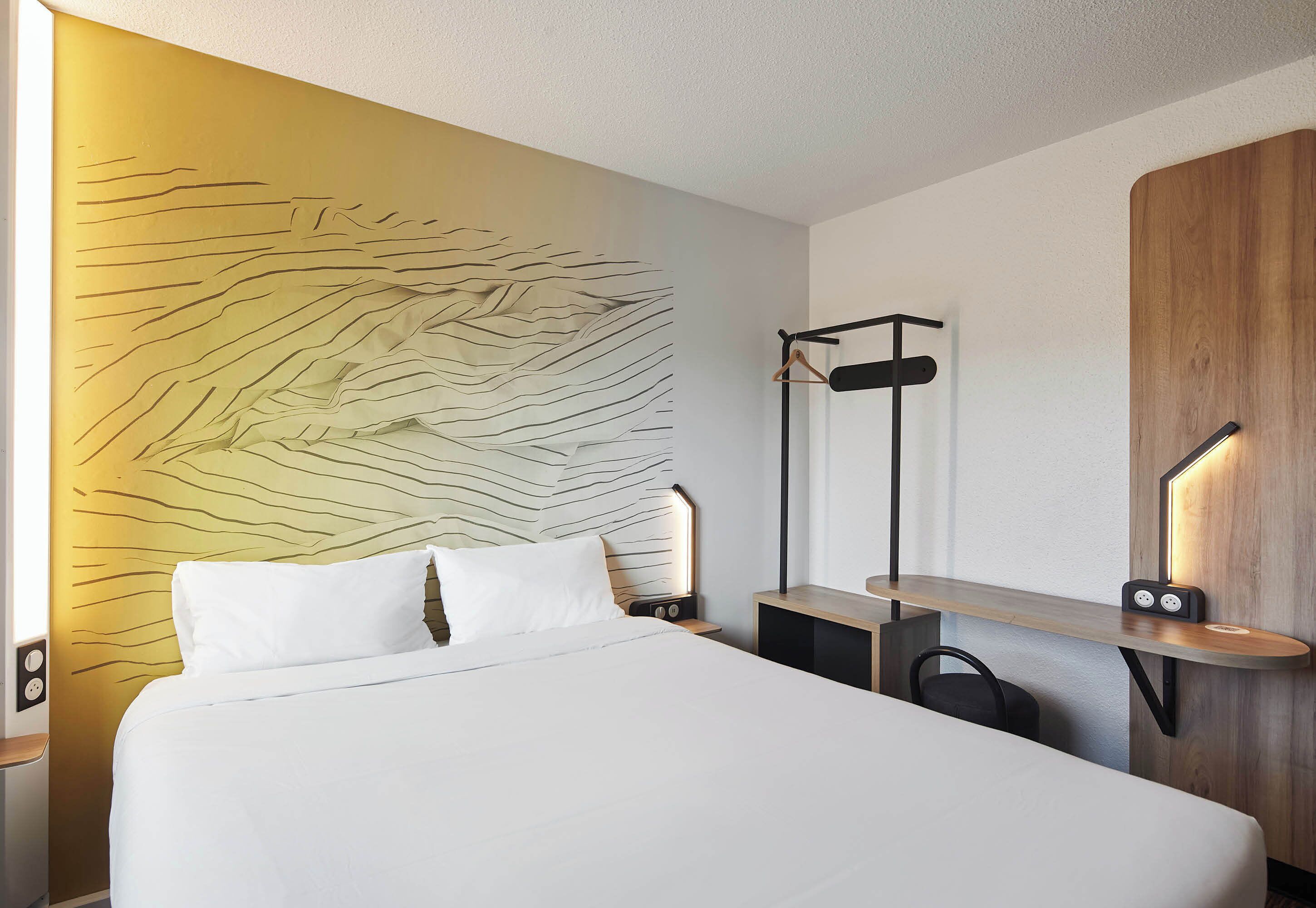A modern hotel room with a large bed, a wooden headboard, a bedside table with a lamp, and a wall-mounted shelf.