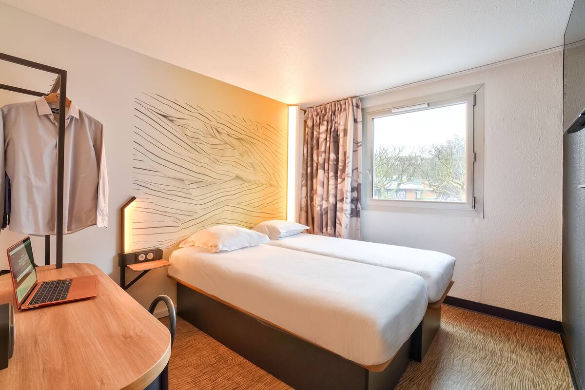 A hotel room with two beds, a desk with a laptop, a wall art piece, and a window with curtains.