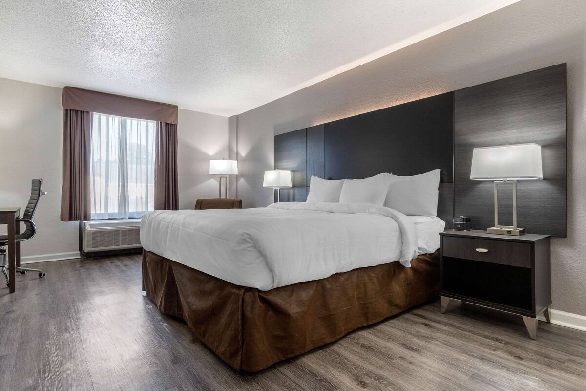 A modern hotel room with a large bed, a desk, and a chair. There is a window with curtains, a bedside table with a lamp, and an air conditioning unit.