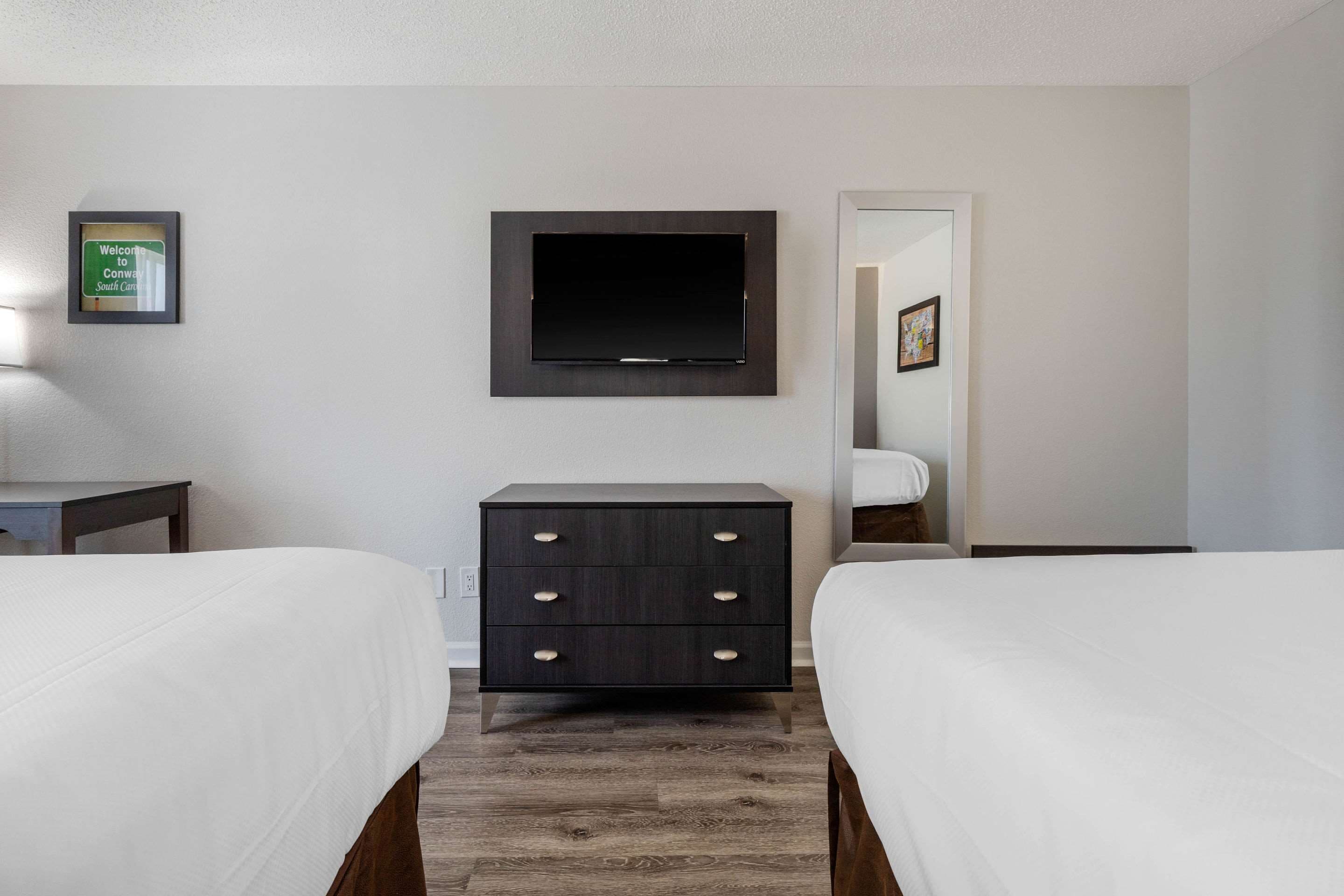 A hotel room with two beds, a flat-screen TV mounted on the wall, a dresser, and a framed welcome sign.