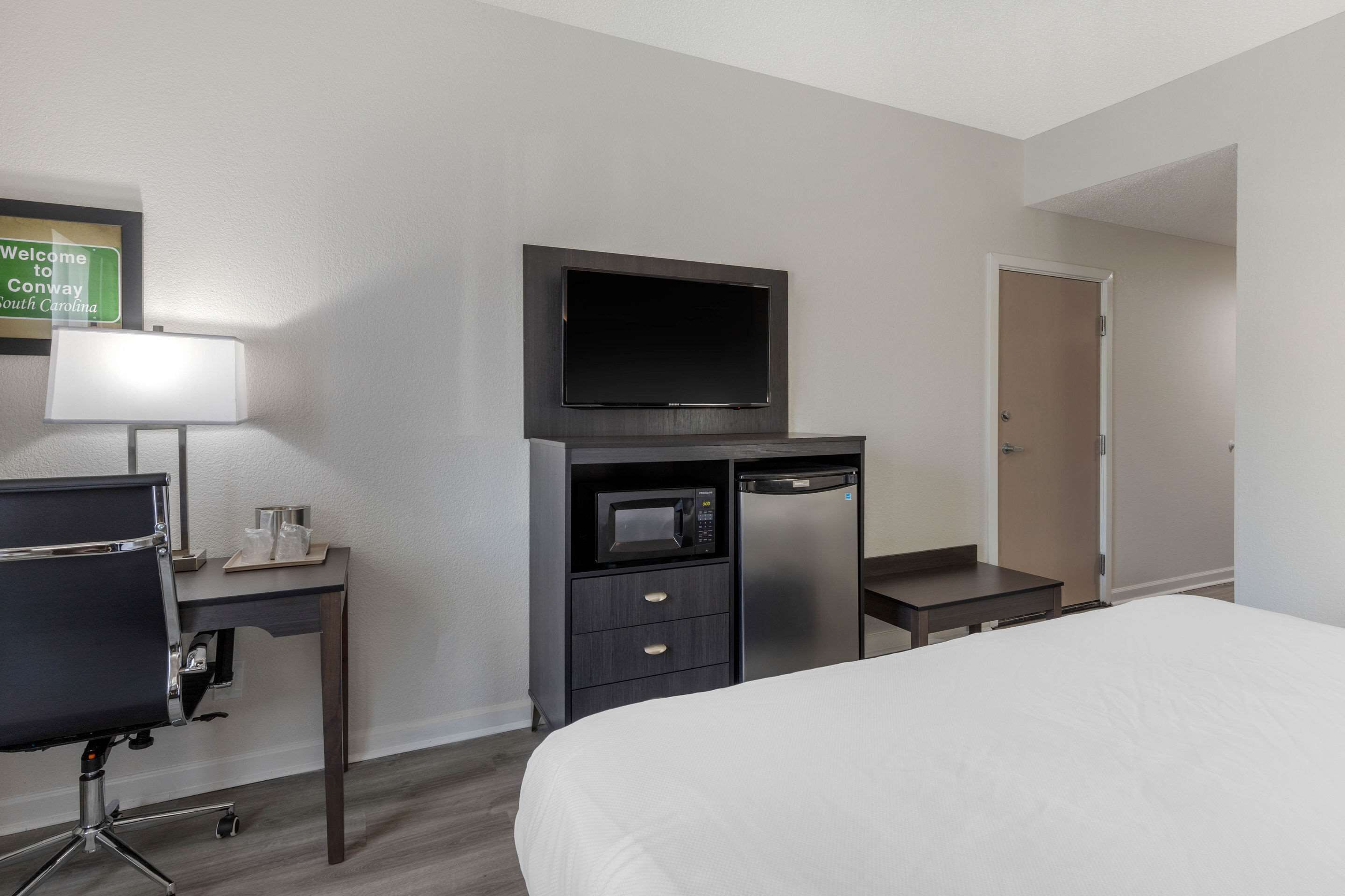 Una habitación de hotel con una cama, un escritorio con computadora, televisor, microondas y puerta.