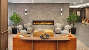 A lobby with a sofa set, a coffee table, two potted plants, a fireplace, and a wall-mounted light fixture.