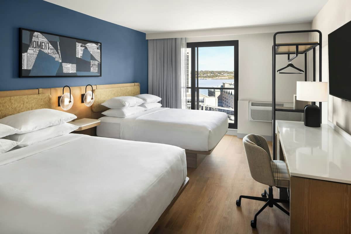 A hotel room with two beds, a desk, a chair, a large window with a view, and a wall-mounted TV.