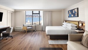 A hotel room with a bed, a desk, a chair, a sofa, and a large window with a view of the sea.