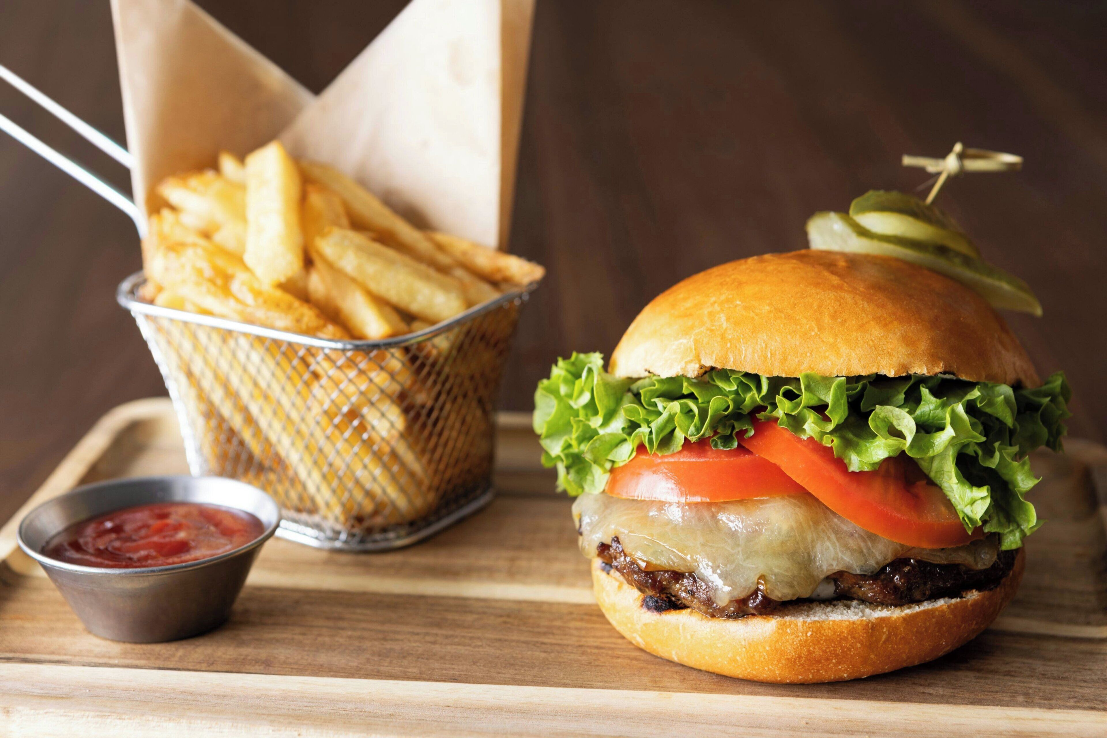 A burger with lettuce, tomato, and cheese, a basket of fries, and a small container of ketchup on a wooden surface.
