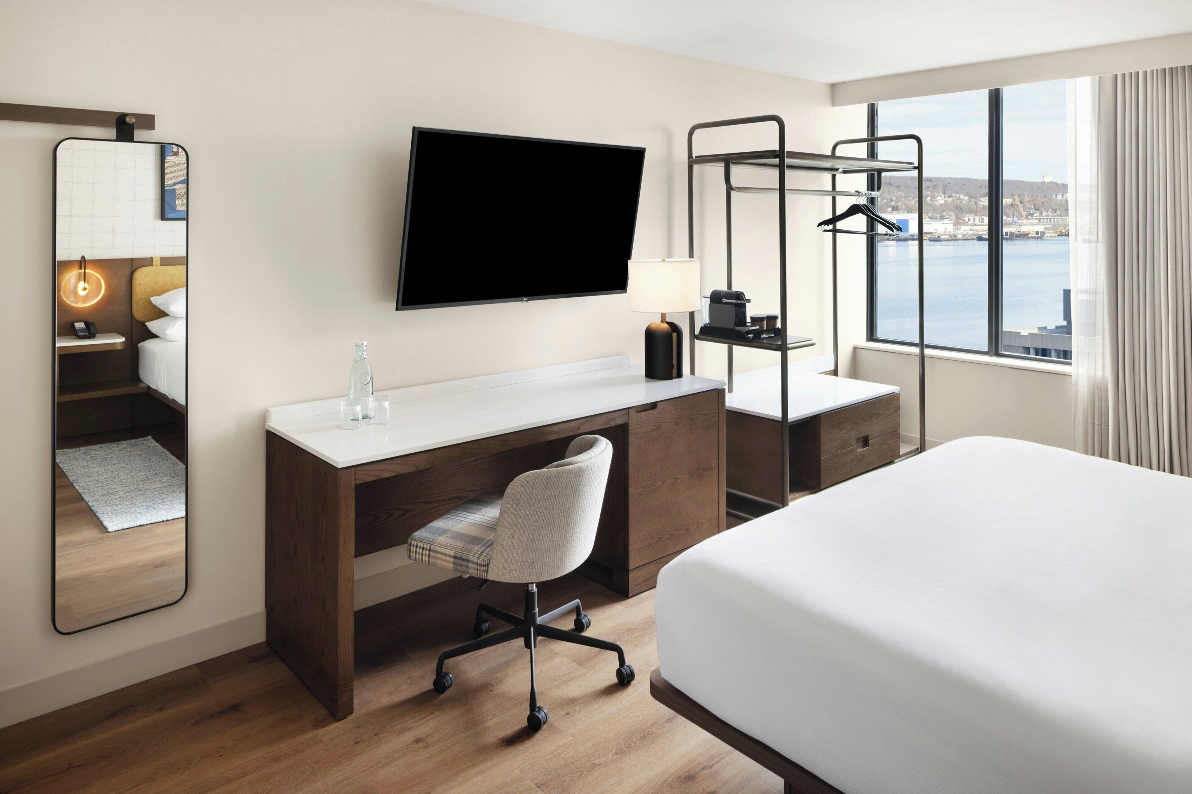 A hotel room with a bed, a desk with a chair, a TV, a mirror, a window with a view, and a hanging rack.